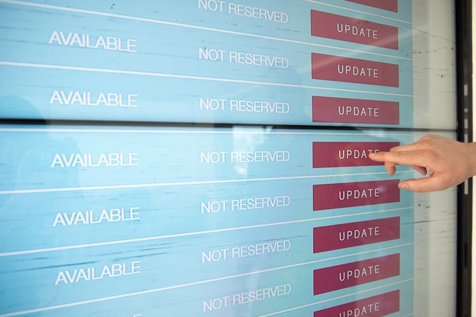 A person's hand is pointing at an electronic booking display with multiple rows showing availability status. The screen displays rows labeled 'AVAILABLE' and 'NOT RESERVED' in white text on a light bl