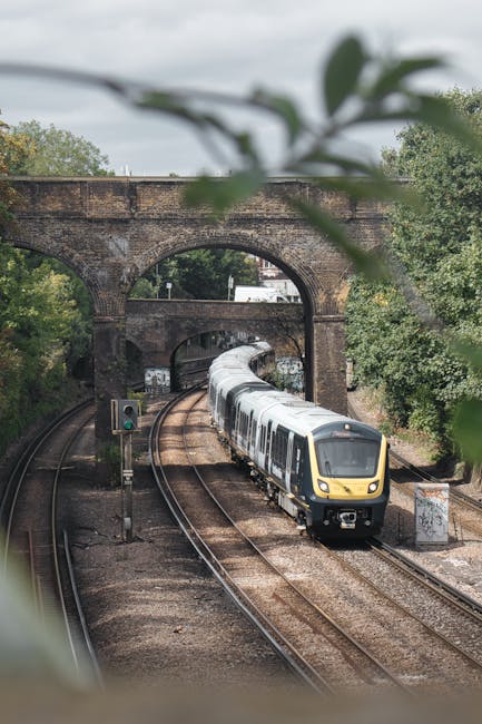The image depicts a quiet railway station platform beneath an overpass bridge on a cloudy day. The platform has a textured yellow safety line along the edge and features a small, empty planter with da